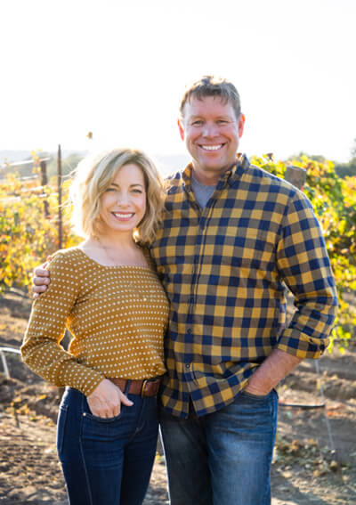 Owners, Bret and Kristen Munselle, are standing together with their arms around each other with a vineyard in the background.