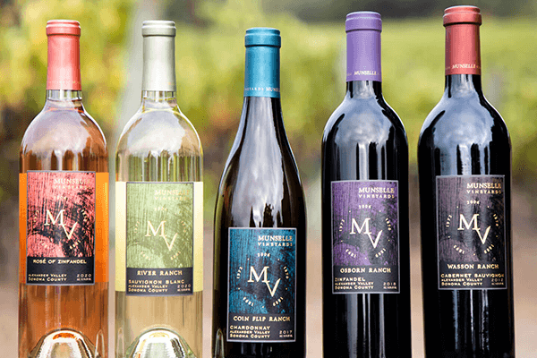 Colorful line up of Munselle Vineyards collection of wines – rosé, sauvignon blanc, chardonnay, zinfandel, and cabernet sauvignon — with a blurred vineyard background.