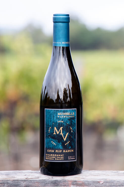 Munselle Vineyards Coin Flip Chardonnay with teal-colored label on a dark glass bottle, with a blurred vineyard background.