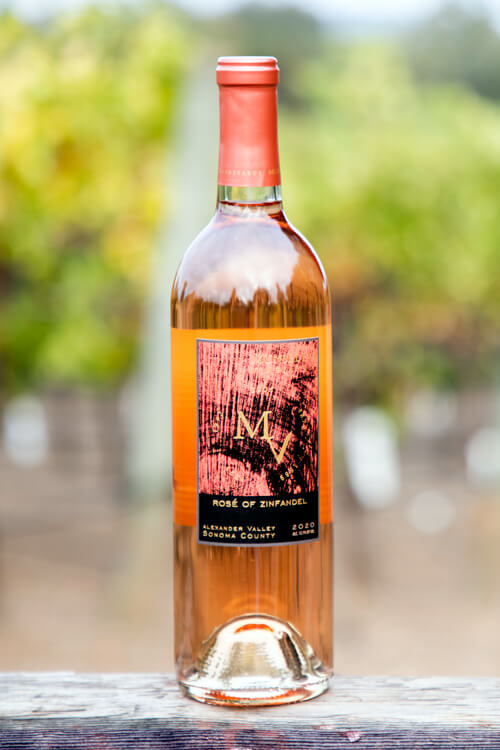 Munselle Vineyards Rose of Zinfandel with rose-colored label on a clear bottle, and a blurred vineyard background.