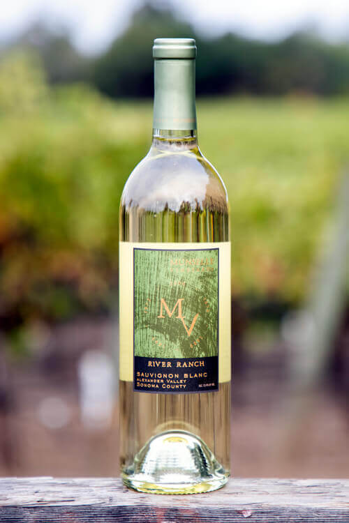 Munselle Vineyards River Ranch Sauvignon Blanc with a light green-colored label on a clear bottle, and a blurred vineyard background.